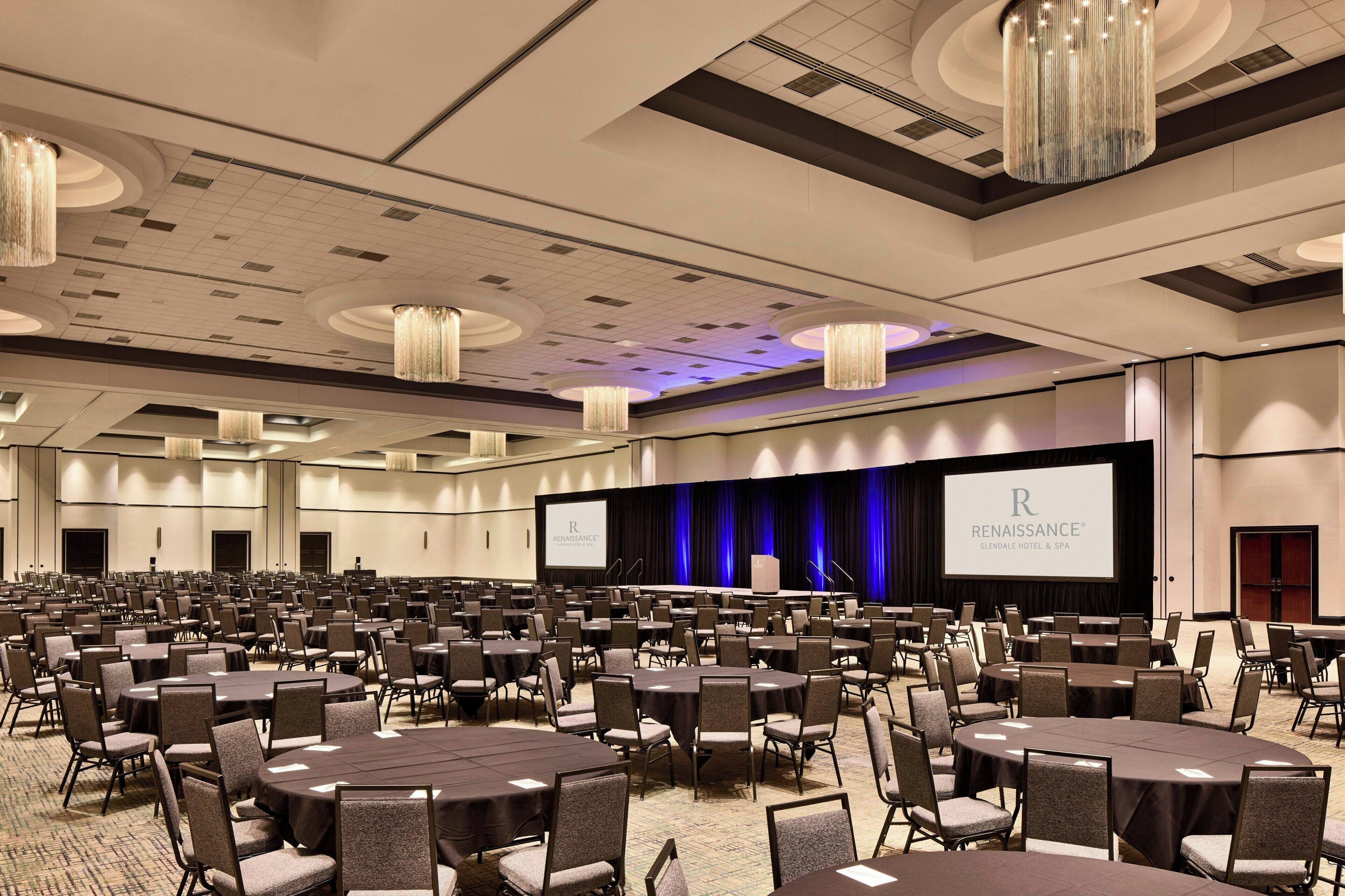A conference room with multiple tables and chairs, a large screen displaying the Renaissance Hotel logo, and a stage with curtains.