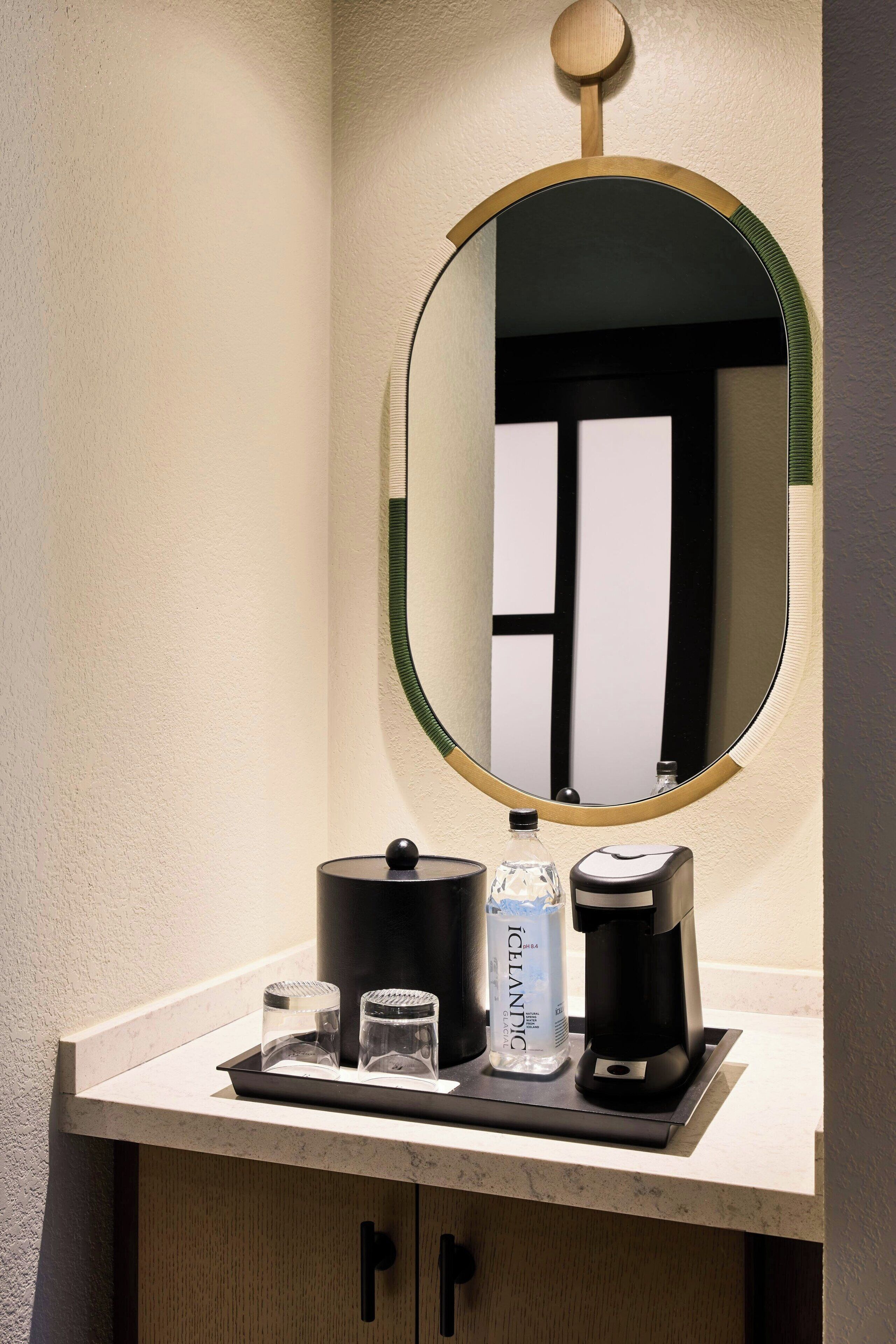 A bathroom vanity with a round mirror, a coffee maker, a bottle of water, and two glasses.