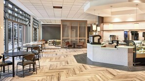 A modern interior with a herringbone wood floor, a reception area with a coffee machine, and large windows offering views of the outside.