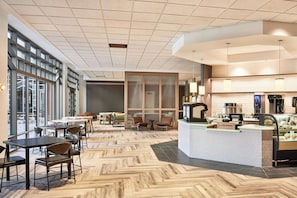 Un intérieur moderne avec un parquet en chevrons, un espace d’accueil doté d’une machine à café et de grandes fenêtres offrant une vue sur l’extérieur.