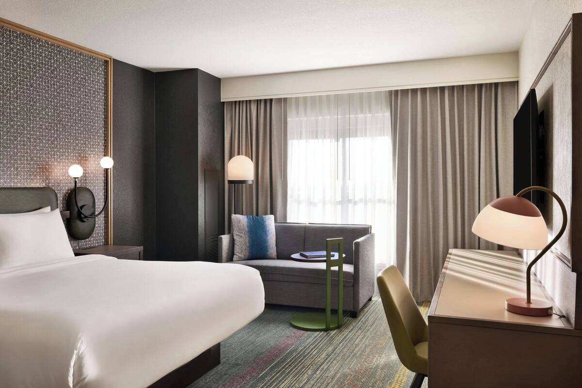 Ein Hotelzimmer mit einem großen Bett, einer Sitzecke, einem Schreibtisch mit Lampe, einem Fernseher und Blick ins Freie durch das Fenster.