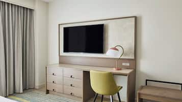 A hotel room with a large flat-screen TV mounted on the wall, a desk with a lamp, a green chair, and a bench.