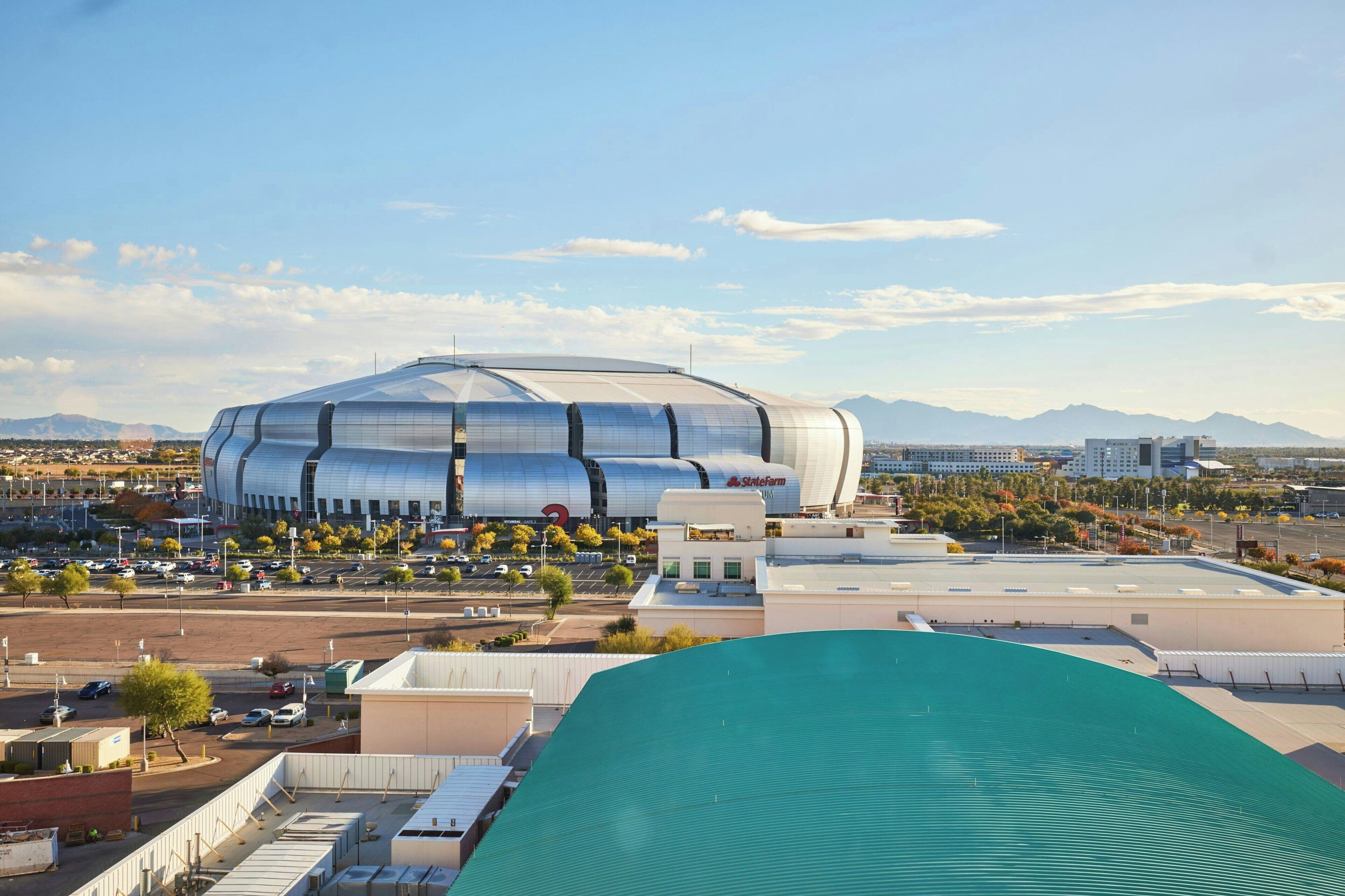 A large, modern stadium with a curved roof, surrounded by a parking lot and urban buildings.