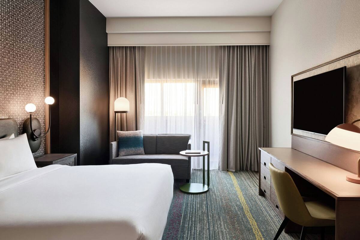 Ein Hotelzimmer mit einem großen Bett, einem Schreibtisch mit Stuhl, einem kleinen Tisch, einer Lampe und einem Fernseher.
