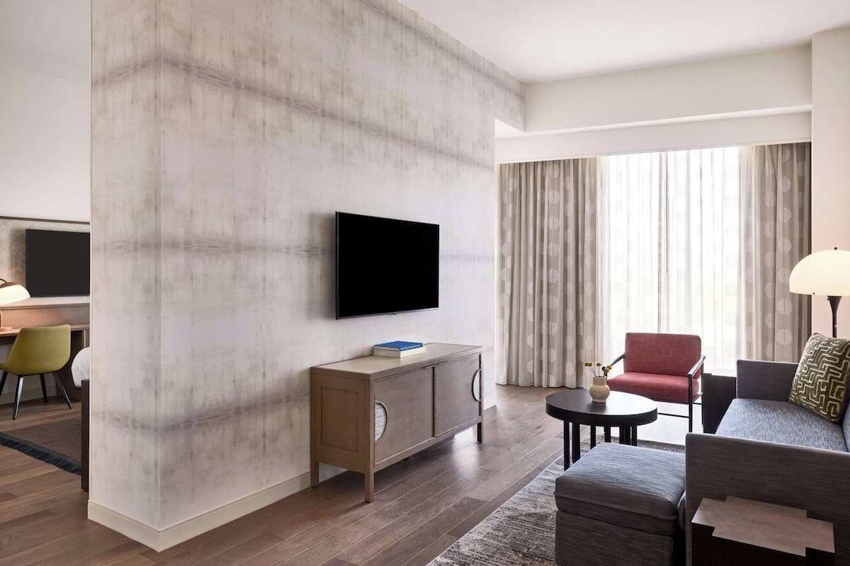 Ein modernes Hotelzimmer mit einem Flachbildfernseher an einer strukturierten Wand, einer Holzkabine mit einem Buch, einem roten Sessel, einem grauen Sofa und einem kleinen runden Tisch mit einer Lampe.