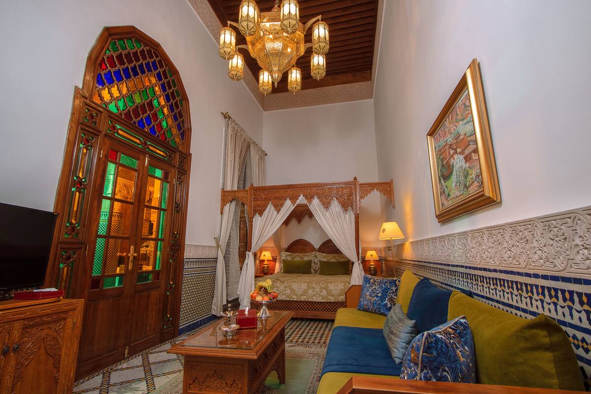 A traditional Moroccan room with a wooden cabinet, a bed with a canopy, a sofa with cushions, and a chandelier.