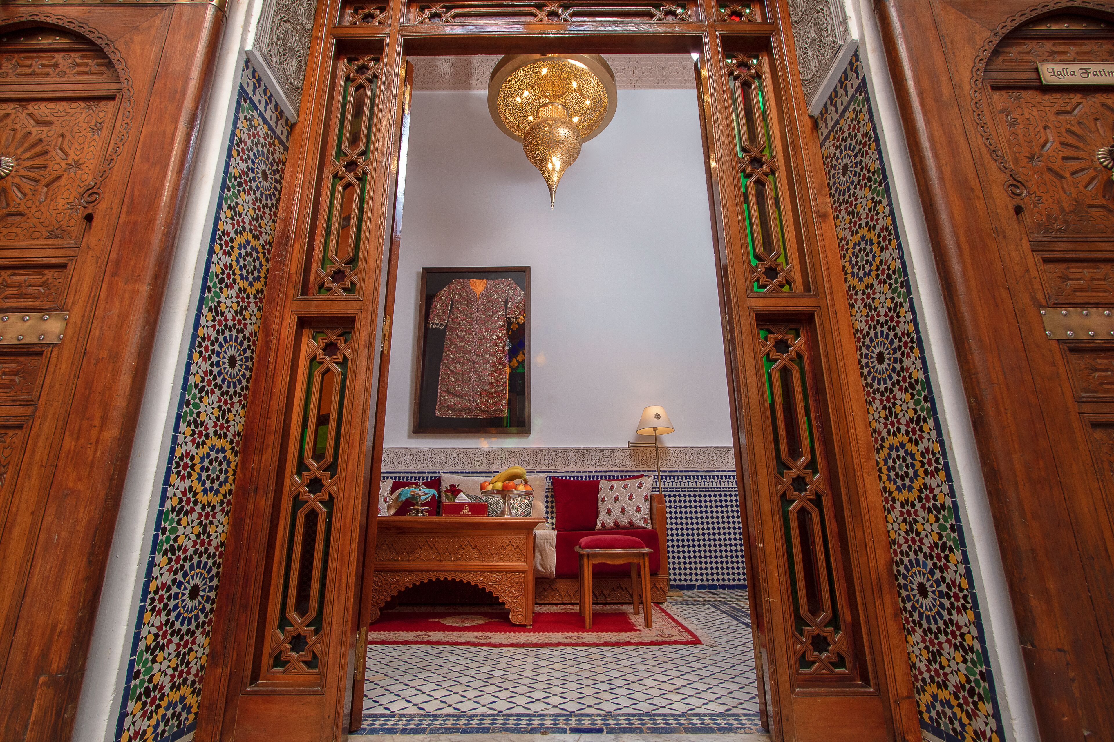 Een klassiek interieur met versierde houten deuren, een vloer met geometrische tegels, een rode bank, een bijzettafeltje met fruit, en een hanglamp.