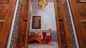 Een klassiek interieur met versierde houten deuren, een vloer met geometrische tegels, een rode bank, een bijzettafeltje met fruit, en een hanglamp.