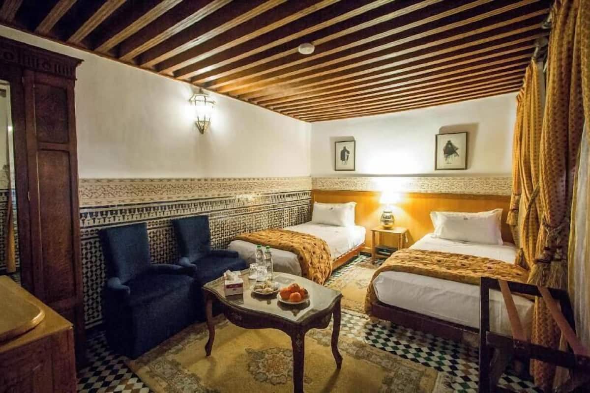 A room with two beds, a sofa, a table with a plate of fruit, and patterned walls.
