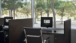 A computer desk with a monitor displaying the Holiday Inn Express logo, situated in a room with large windows overlooking a tree-lined street. - Holiday Inn Express Dubai, Internet City by IHG (Dubai)