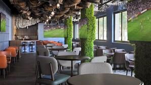 A restaurant with round tables and chairs, a bar area, and large windows with a view of a soccer field. - Holiday Inn Express Dubai, Internet City by IHG (Dubai)
