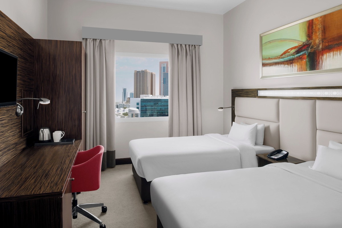 Quarto de hotel com duas camas, uma escrivaninha, uma cadeira, um telefone e uma janela com vista para a cidade.