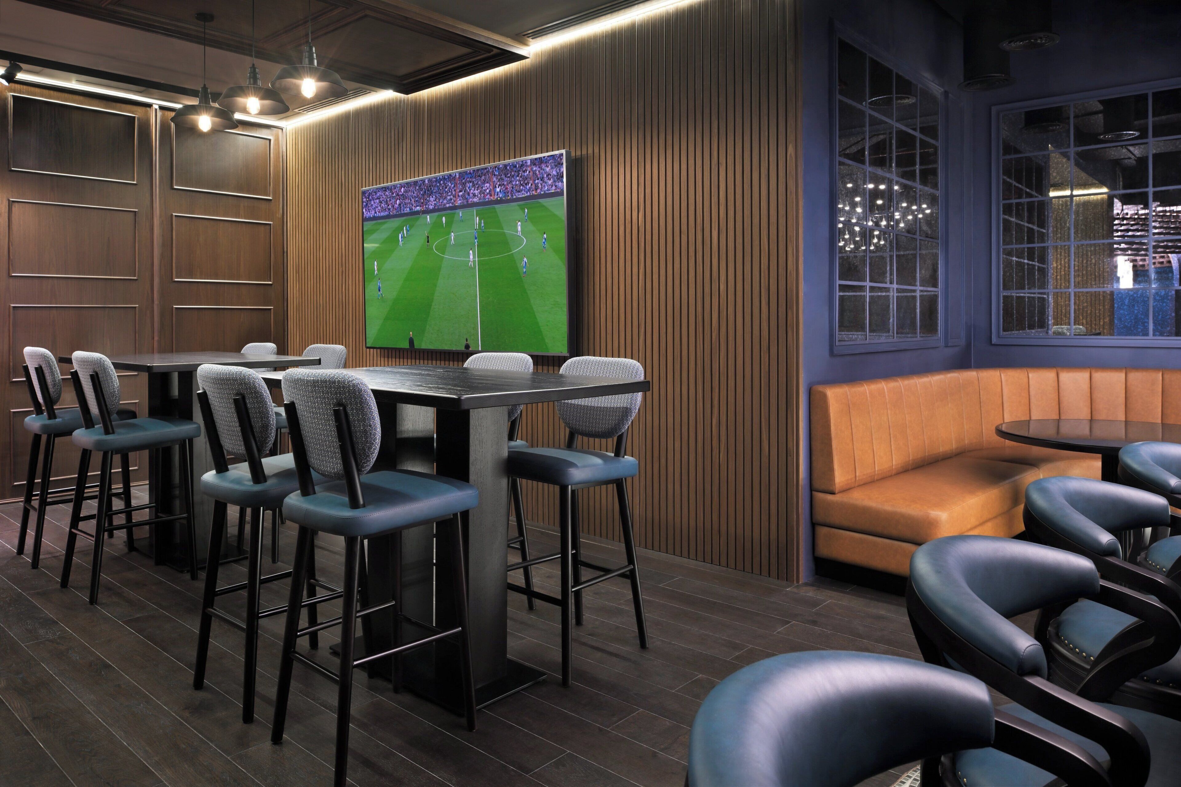 Un espace lounge moderne doté d’un grand écran de télévision diffusant un match de football, de lambris en bois et d’un mélange de sièges de bar et de banquettes.