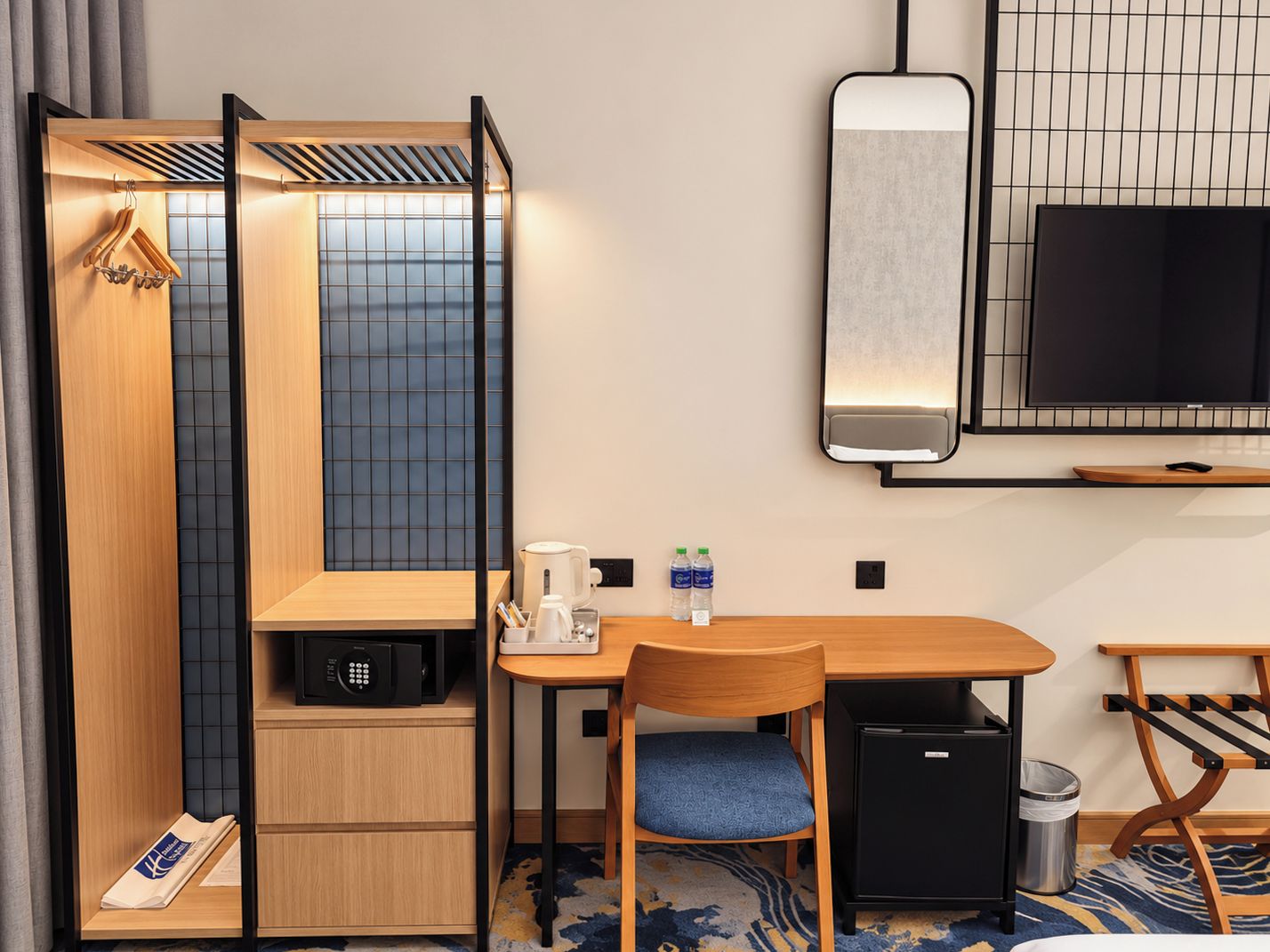 A compact hotel room with a bed, a desk, a chair, a microwave, a small refrigerator, a TV, and a wall-mounted heater.
