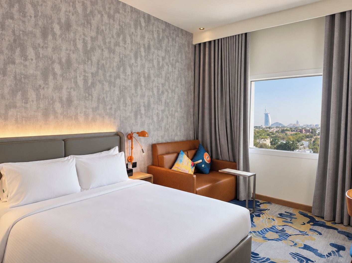 Una habitación de hotel con una cama grande, una mesita de noche, un área de descanso con cojines, una ventana con cortinas y vistas a un paisaje urbano.
