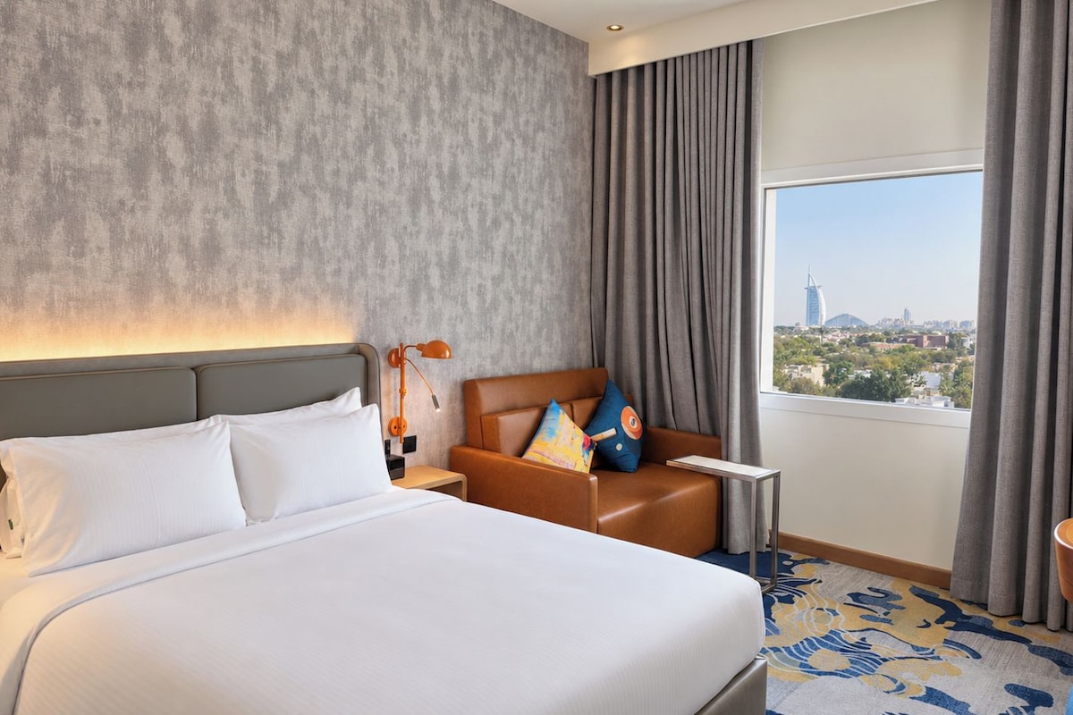 A hotel room with a large bed, a nightstand, a seating area with cushions, a window with curtains, and a view of a cityscape.