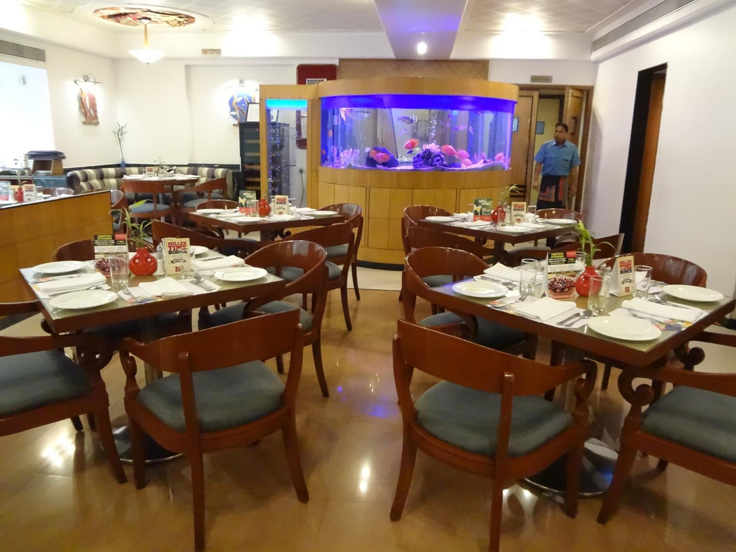 Un restaurant avec des tables et des chaises en bois, un grand aquarium au centre et une personne debout à l’arrière-plan.