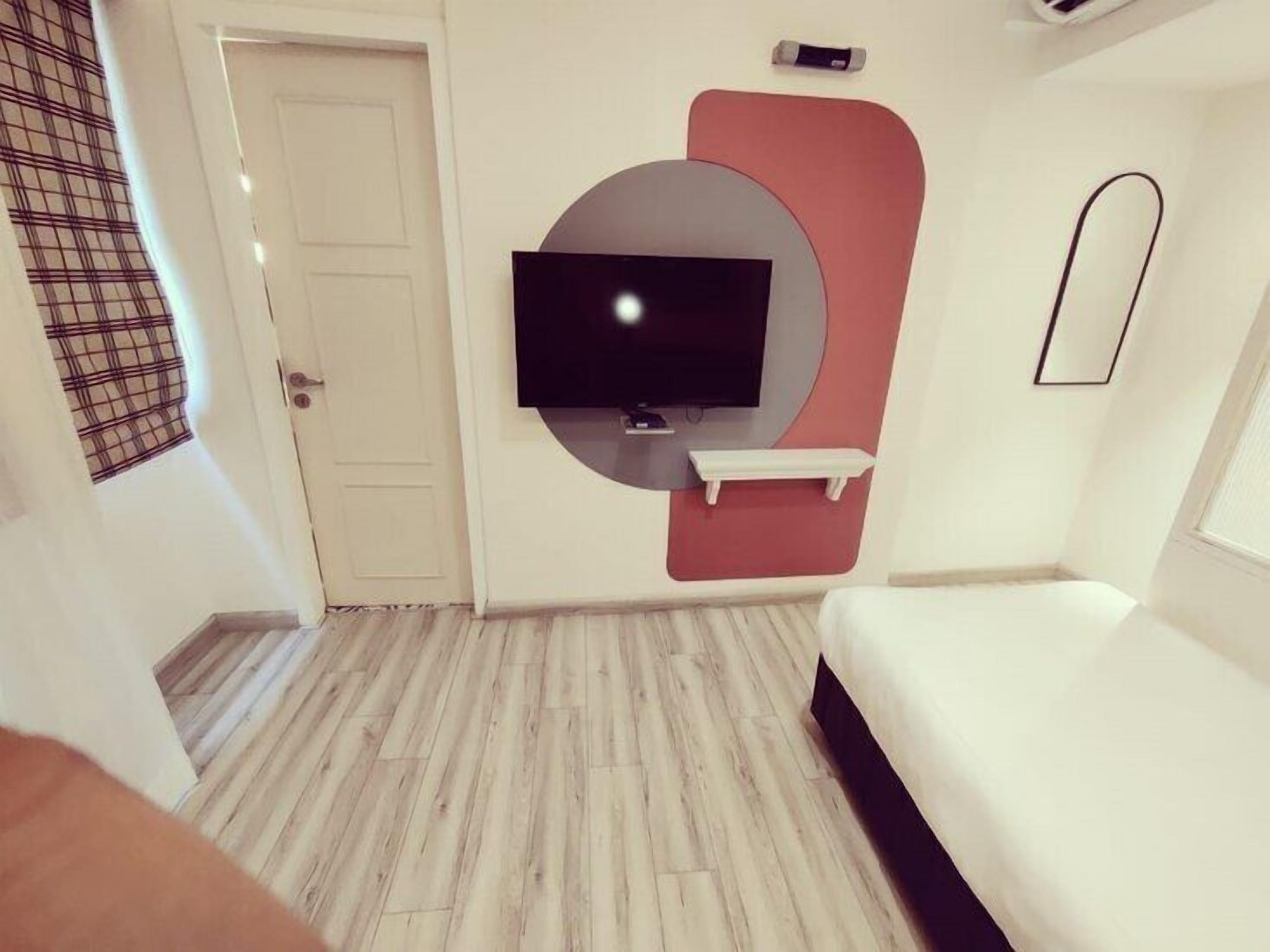 A modern hotel room with a flat-screen TV mounted on a wall, a small white shelf below it, and a bed with a dark headboard.
