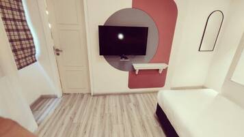 Una habitación de hotel moderna con un televisor de pantalla plana montado en la pared, un pequeño estante blanco debajo y una cama con cabecera oscura.