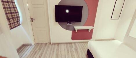 A modern hotel room with a flat-screen TV mounted on a wall, a small white shelf below it, and a bed with a dark headboard.