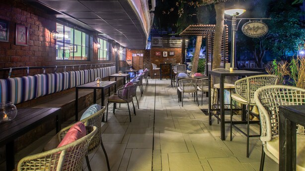 An outdoor restaurant with wooden tables and wicker chairs, a brick wall with framed pictures, and a Red Bull sign. - T24 Retro (Mumbai)