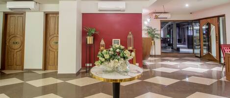 Un vestíbulo de hotel con suelo a cuadros, una mesa redonda central con un jarrón de flores, puertas de madera con letreros de 'L' y 'R', y una pared de acento roja.