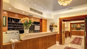 A reception area with a wooden counter, a phone, a vase with flowers, a wine cooler, and a bar in the background. - Hotel San Marco (Rome)