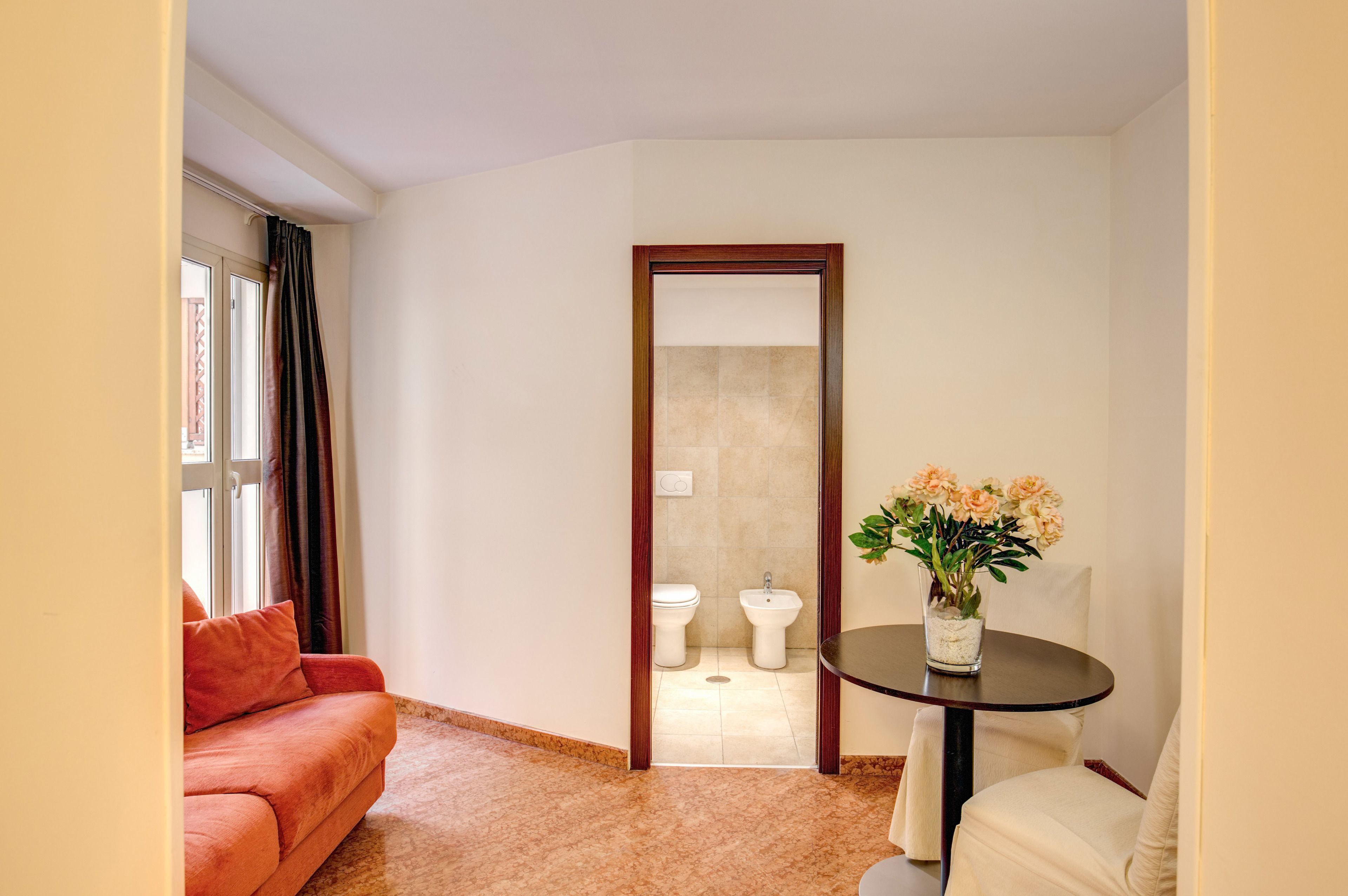 A hotel room with a red sofa, a round table with a vase of flowers, a mirror, and a view of a bathroom with a toilet and bidet.