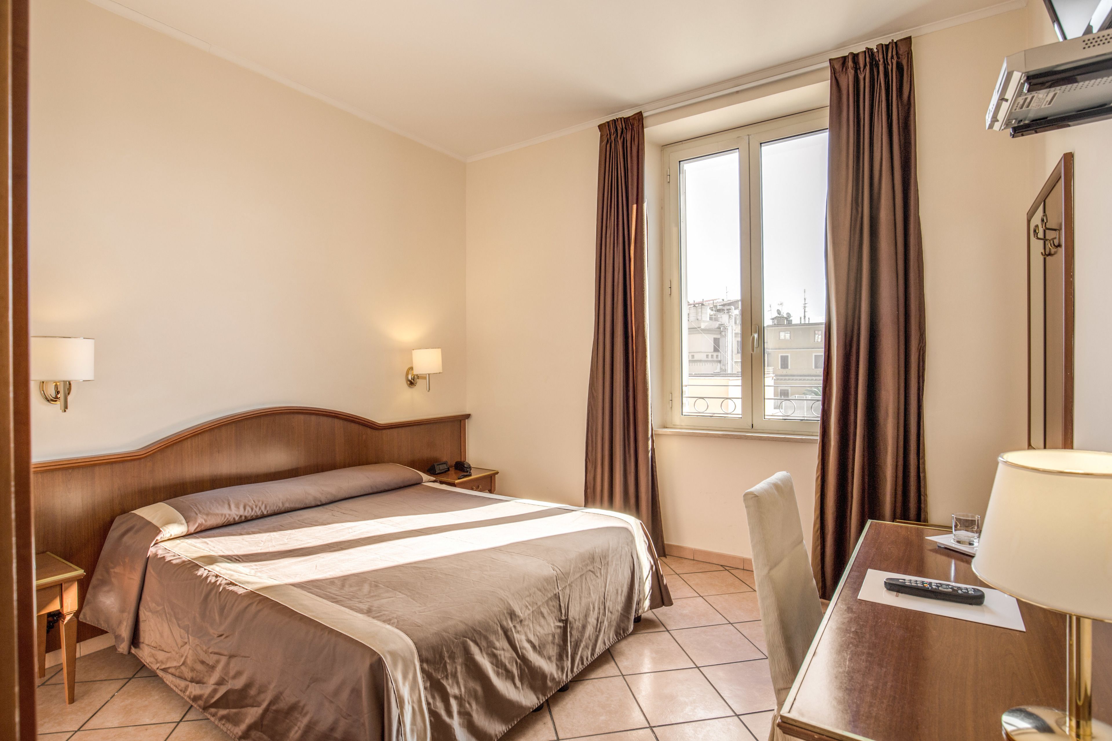 Una habitación de hotel con cama, mesitas de noche, un escritorio con lámpara y una ventana con cortinas.