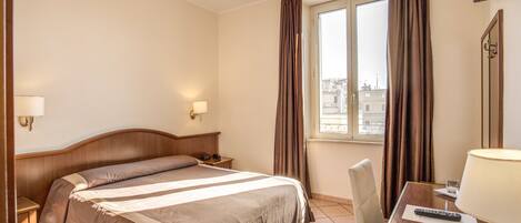 Una habitación de hotel con cama, mesitas de noche, un escritorio con lámpara y una ventana con cortinas.