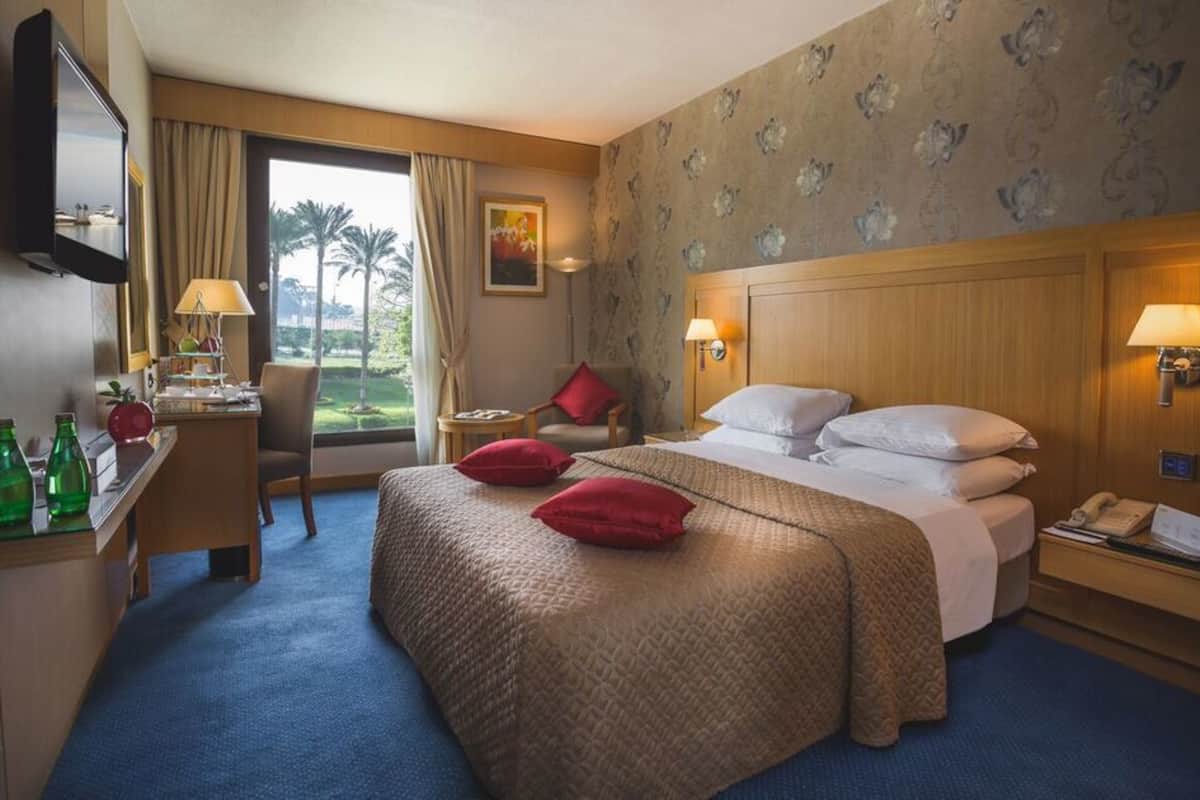 Ein Hotelzimmer mit einem großen Bett, einem Schreibtisch mit Lampe, einem Sessel und Blick auf Palmen durch das Fenster.
