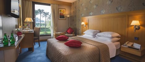 A hotel room with a large bed, a desk with a lamp, a chair, and a view of palm trees through the window.