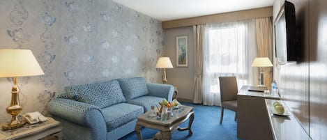 A hotel room with a blue patterned sofa, a glass coffee table, a desk with a flat-screen TV, and two lamps.