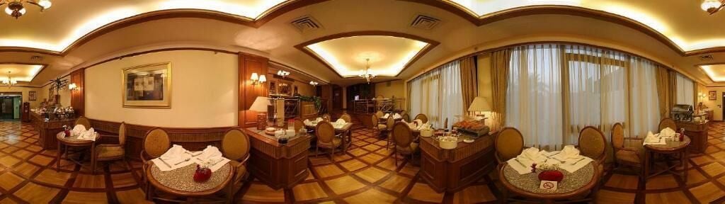 A panoramic view of a hotel dining area with multiple tables and chairs, a bar, and a reception desk.