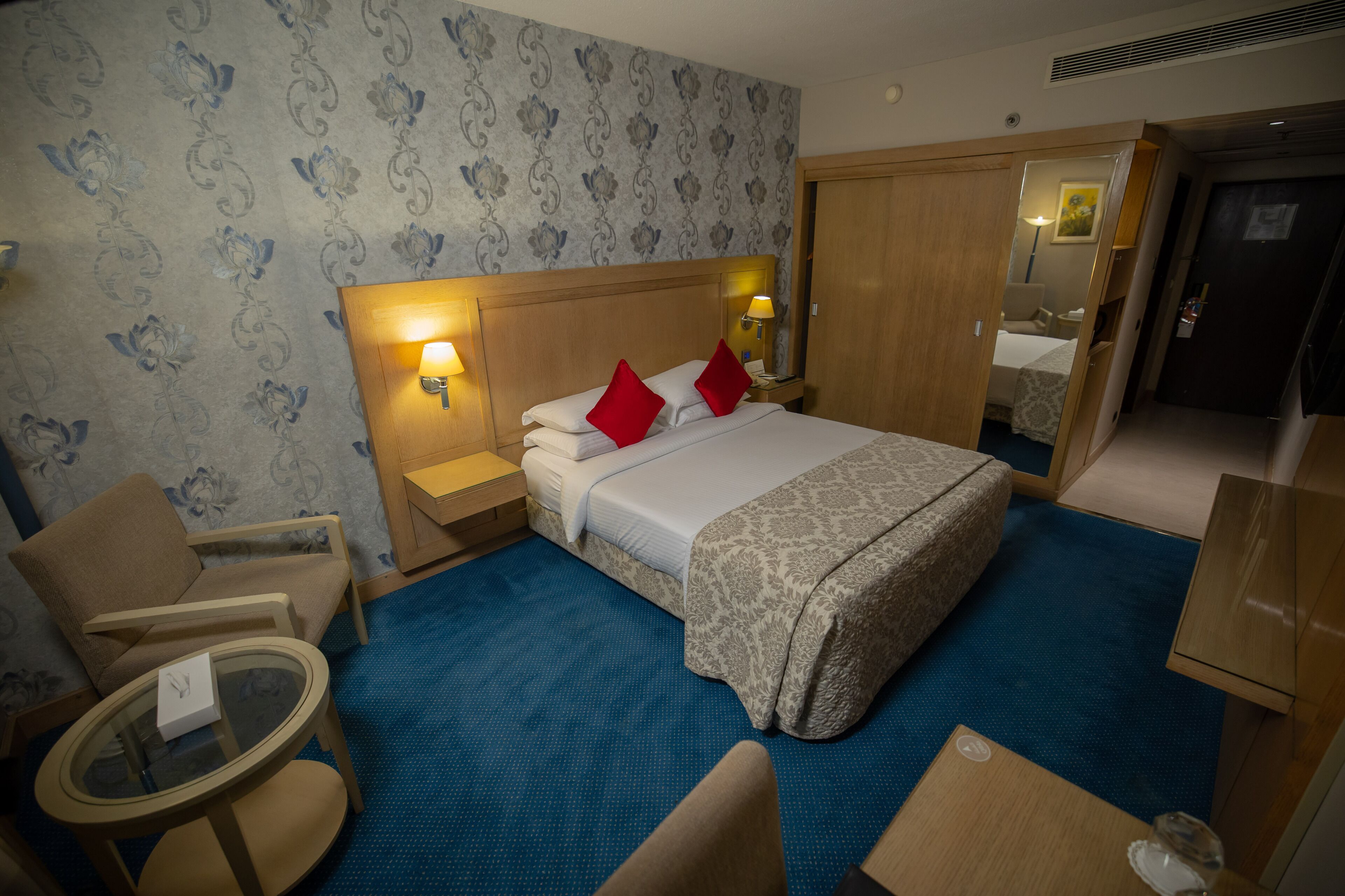 A hotel room with a bed, two nightstands with lamps, a round table, a chair, a wardrobe, and a mirror.
