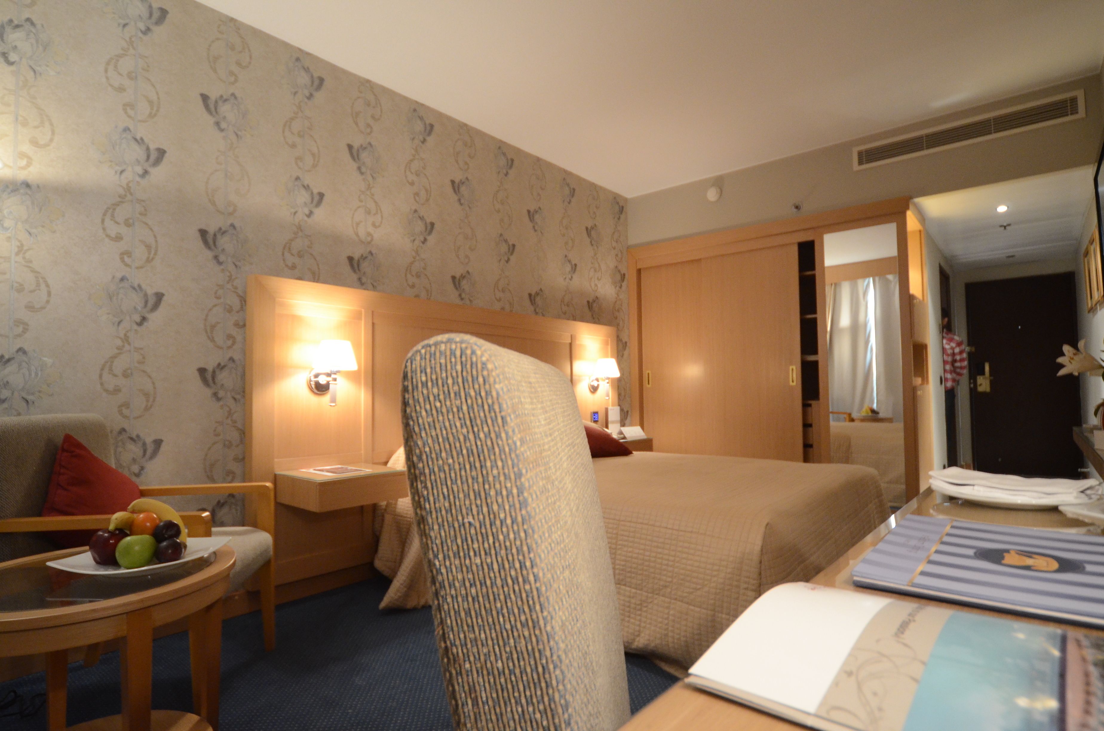 A hotel room with a bed, a chair, a table with fruits, and a bedside table with a lamp.
