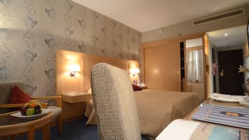 A hotel room with a bed, a chair, a table with fruits, and a bedside table with a lamp.