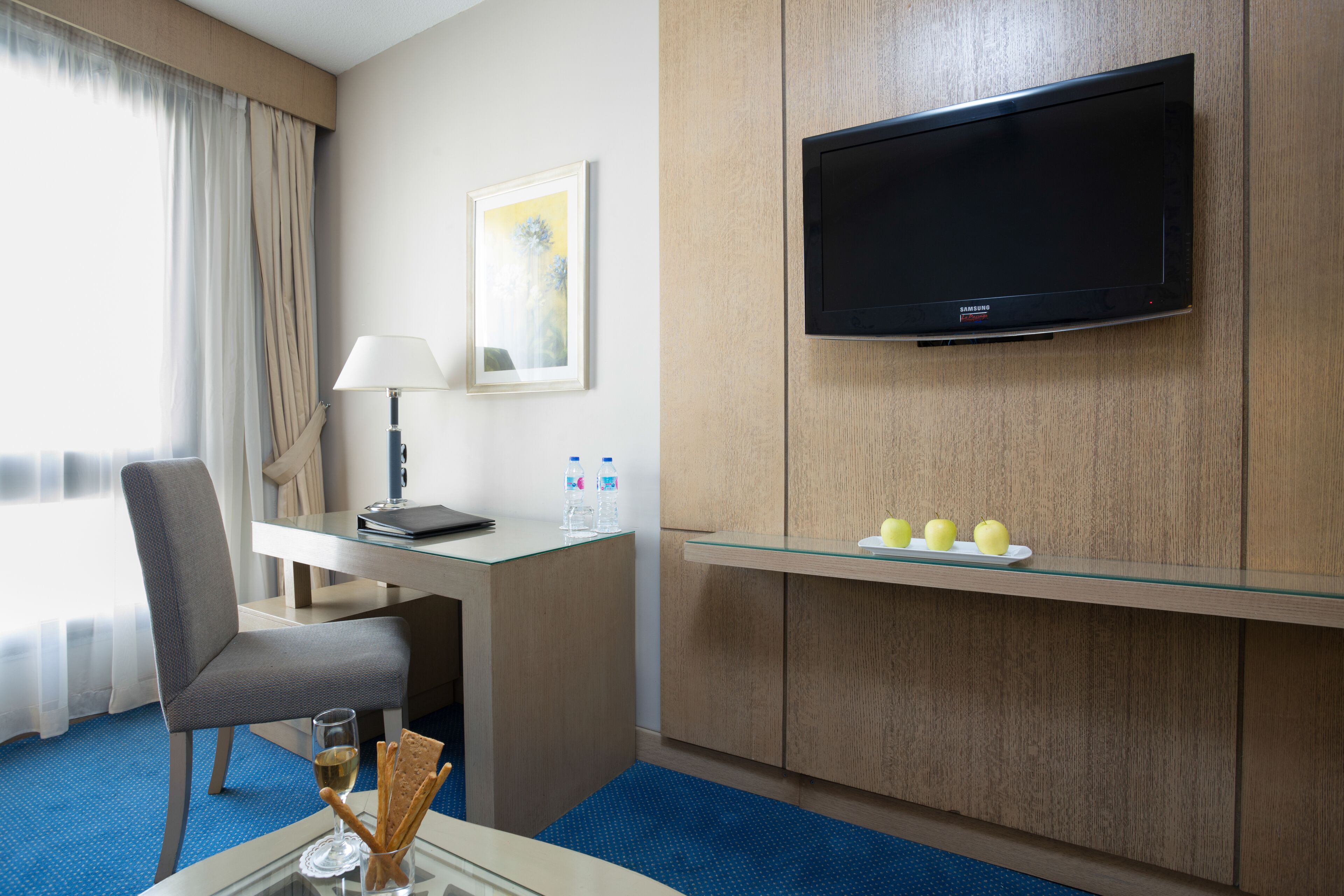 A hotel room with a desk, chair, flat-screen TV, and a glass shelf with apples.