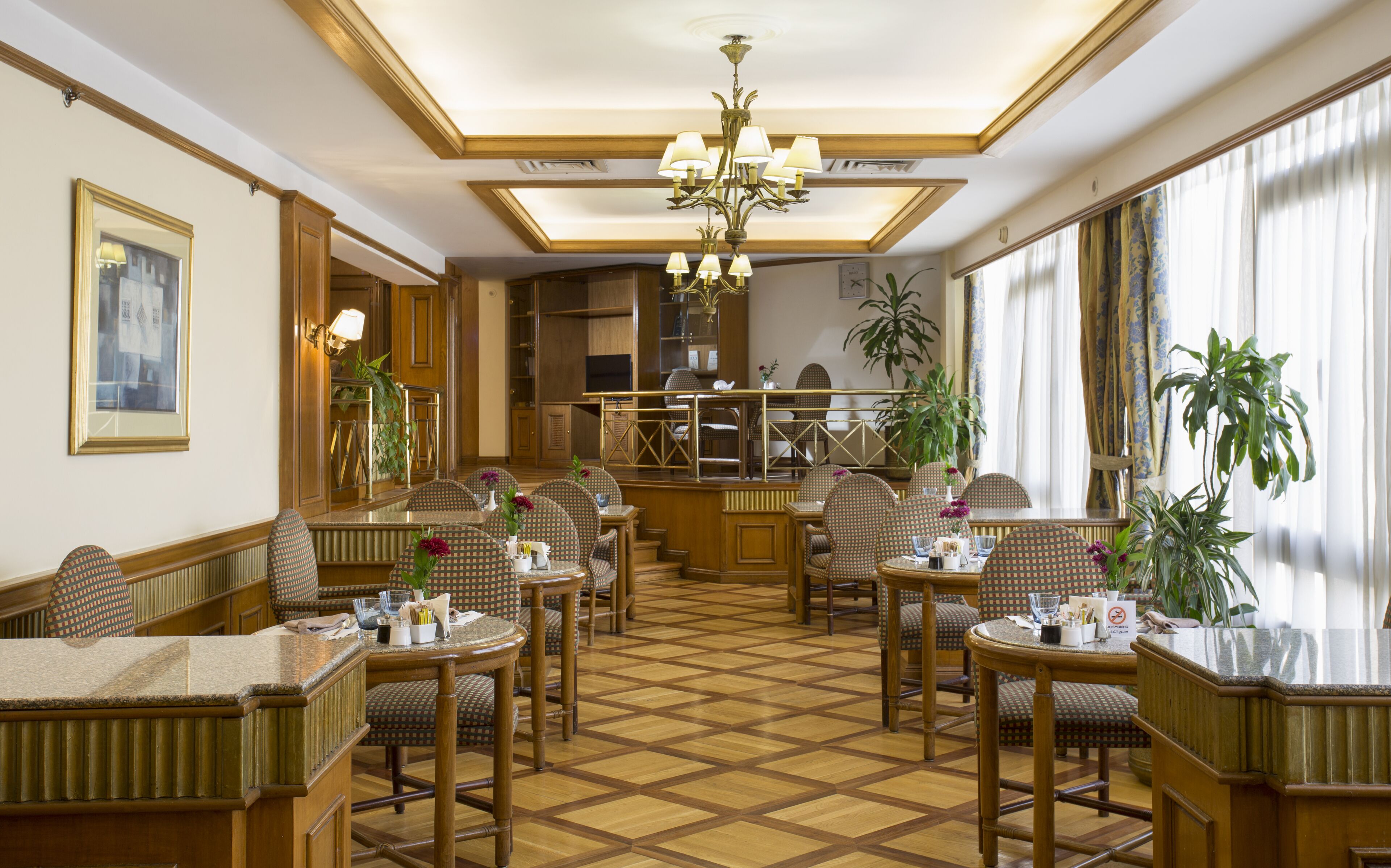 Un hall d’hôtel avec un sol à carreaux, des meubles en bois, un lustre et de grandes fenêtres avec des rideaux.