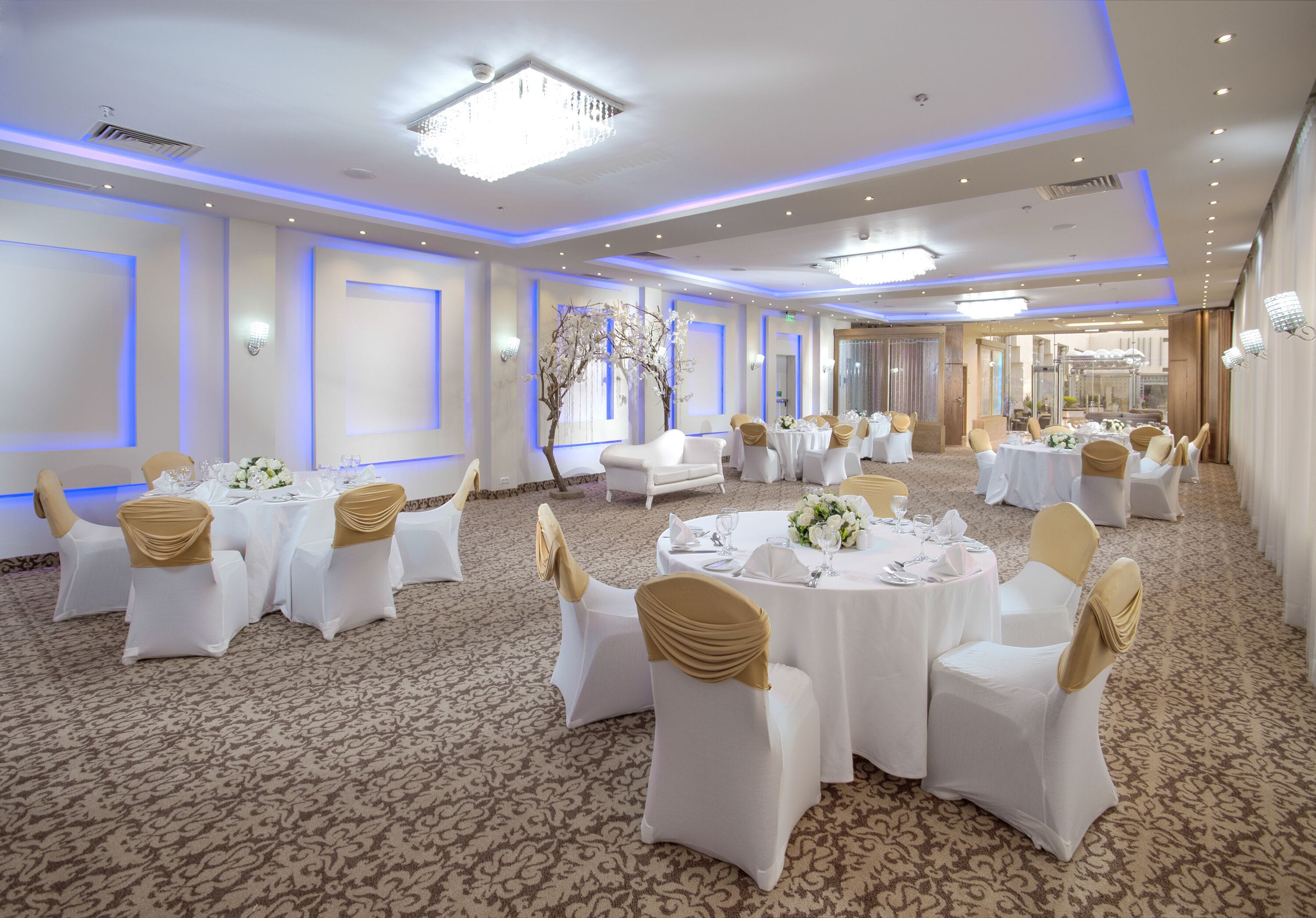A banquet hall with round tables, white chairs with gold sashes, and a carpeted floor.