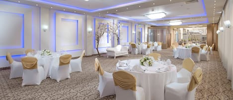 A banquet hall with round tables, white chairs with gold sashes, and a carpeted floor.