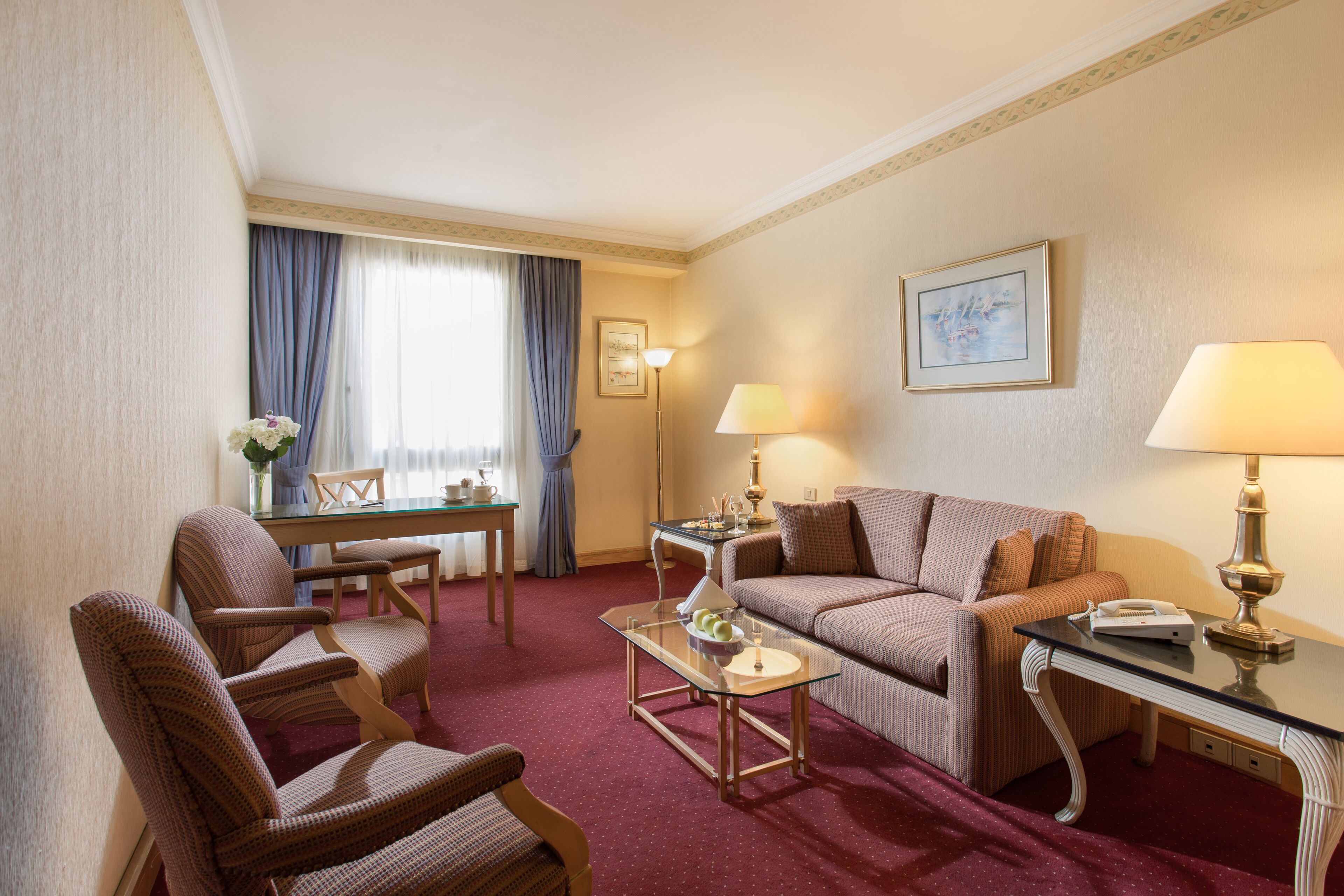 A hotel room with a sofa, armchairs, a glass coffee table, a desk, and lamps.