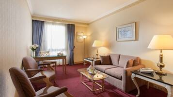 A hotel room with a sofa, armchairs, a glass coffee table, a desk, and lamps.