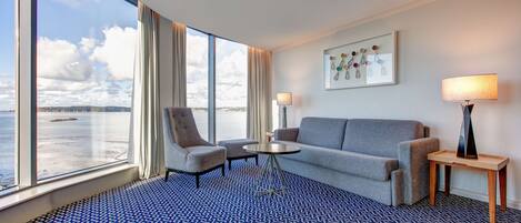 Habitación de hotel con un ventanal grande, un sofá, dos sillones, una mesita y una lámpara.