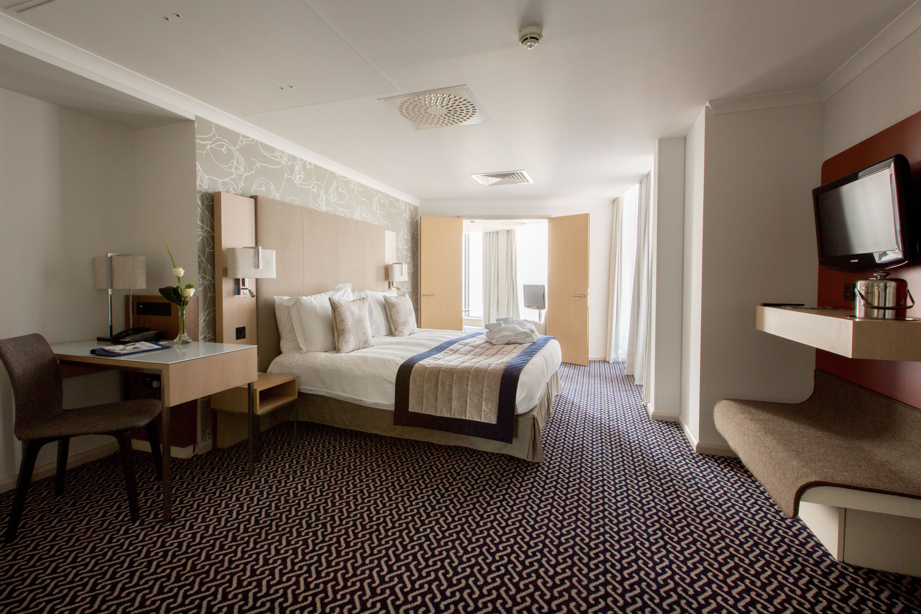 A hotel room with a bed, desk, chair, TV, and a patterned carpet.