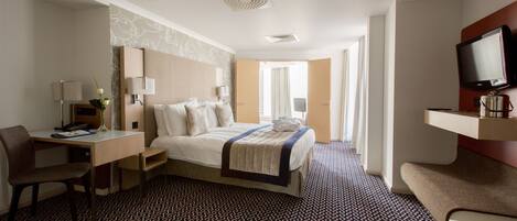 A hotel room with a bed, desk, chair, TV, and a patterned carpet.