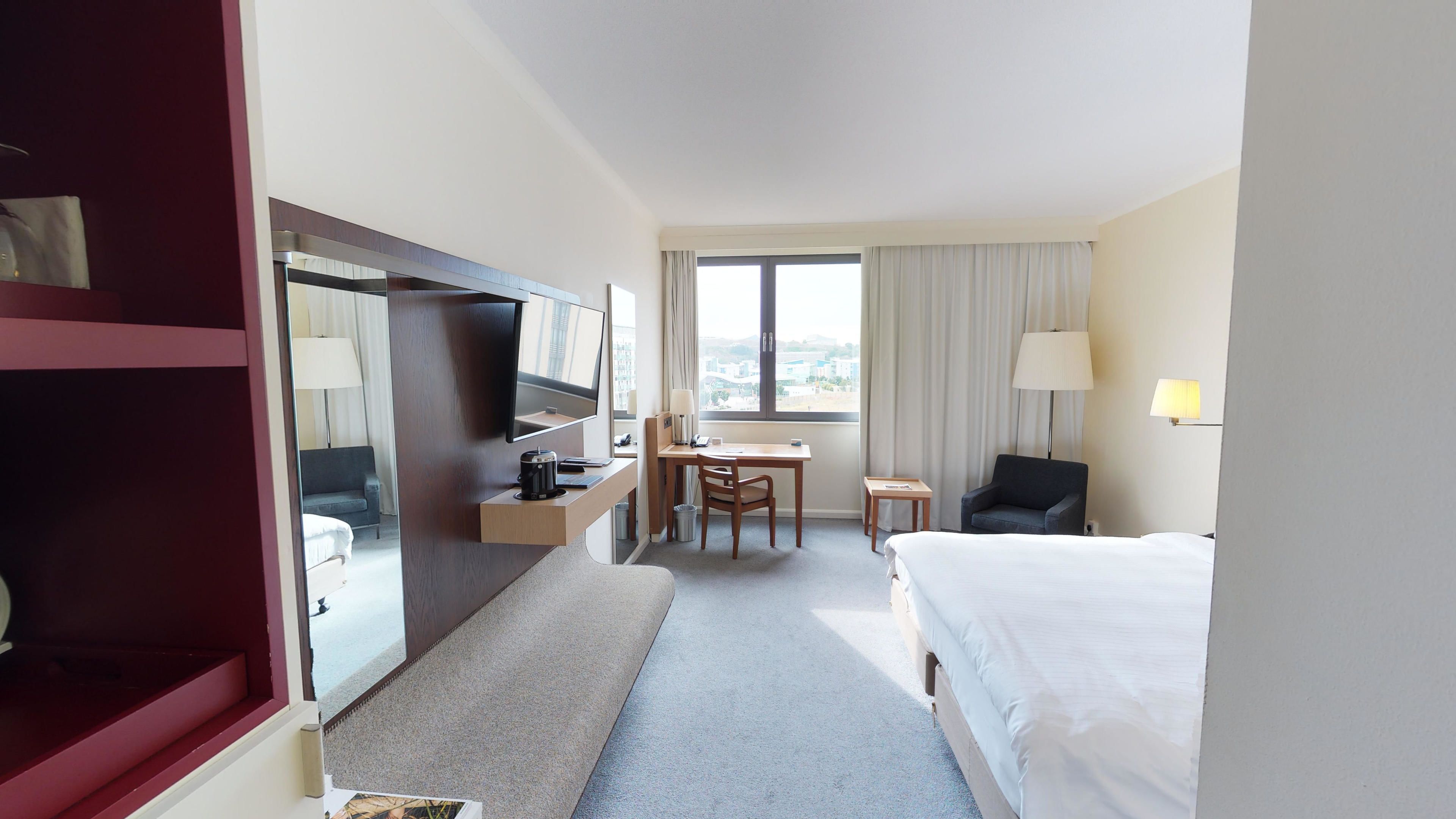 A hotel room with a bed, a desk with a chair, a TV, a lamp, and a window with curtains.