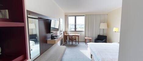 A hotel room with a bed, a desk with a chair, a TV, a lamp, and a window with curtains.