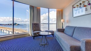 Quarto de hotel com uma janela ampla que oferece vista para uma marina e para a água. Há um sofá, uma cadeira e uma pequena mesa redonda.