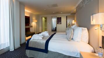 A hotel room with a neatly made bed, a bedside table with a bottle of water, a mirror, and a wall-mounted TV.